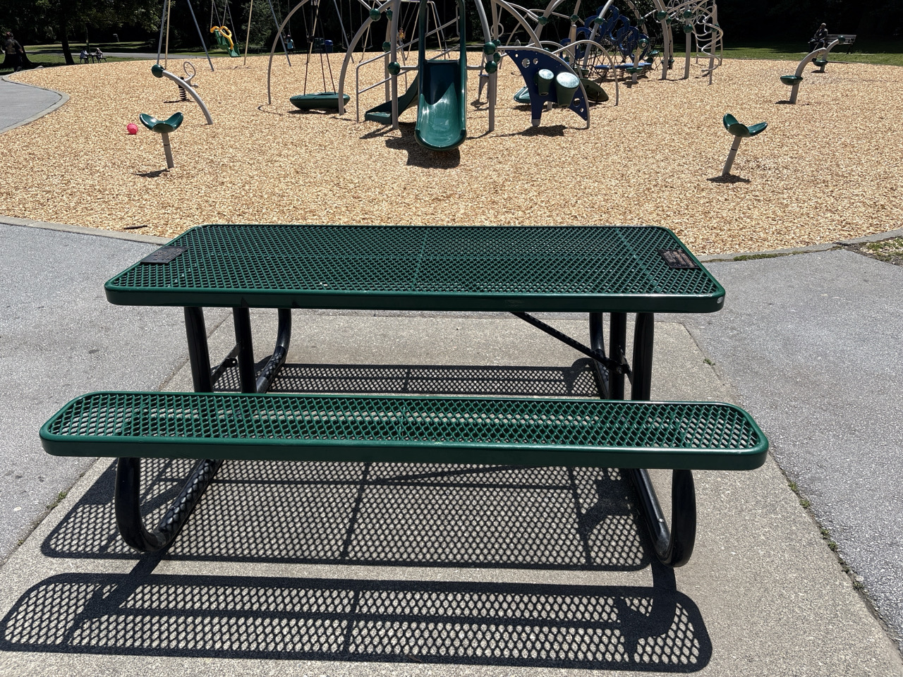 Green metal picnic table on a paved surface beside a playground with play equipment in the background.