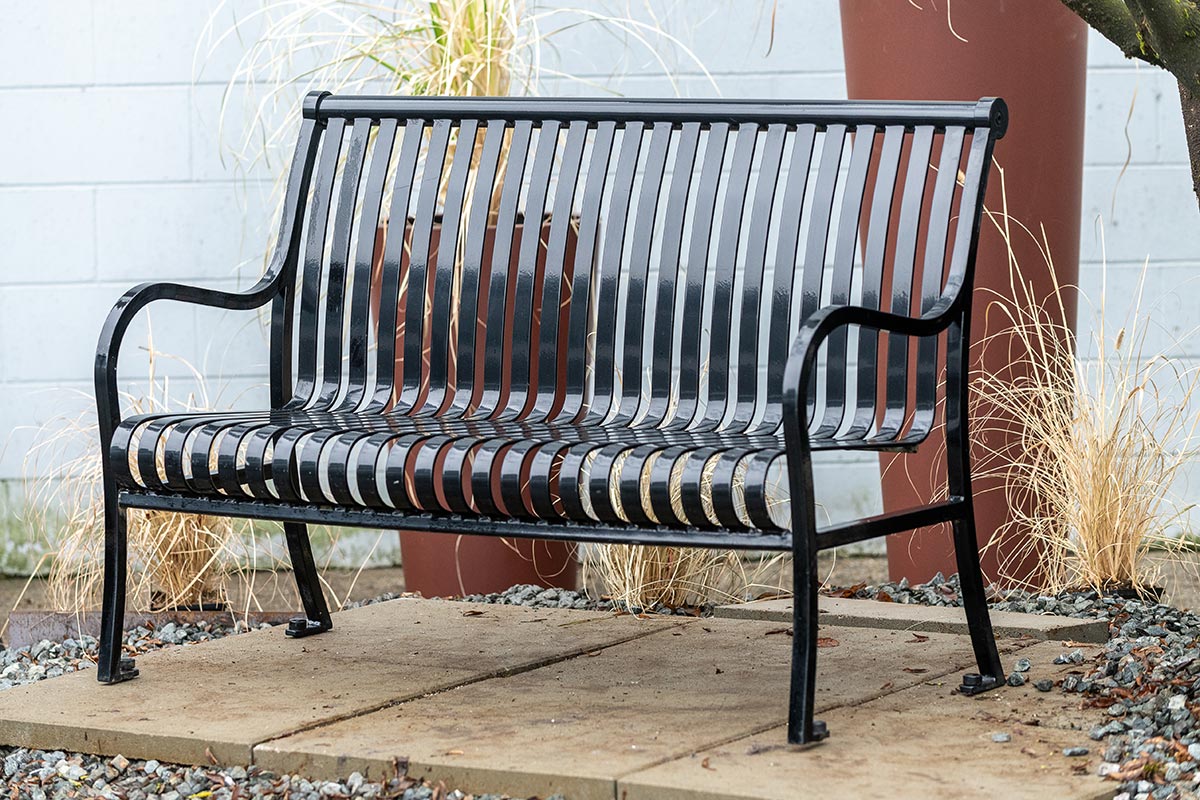 Black metal park bench on a concrete pad surrounded by ornamental grasses and landscaping.