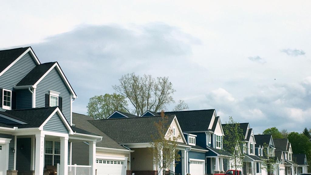 Row of modern suburban homes with dark roofs and blue siding, featuring front porches and garages, under a partly cloudy sky.