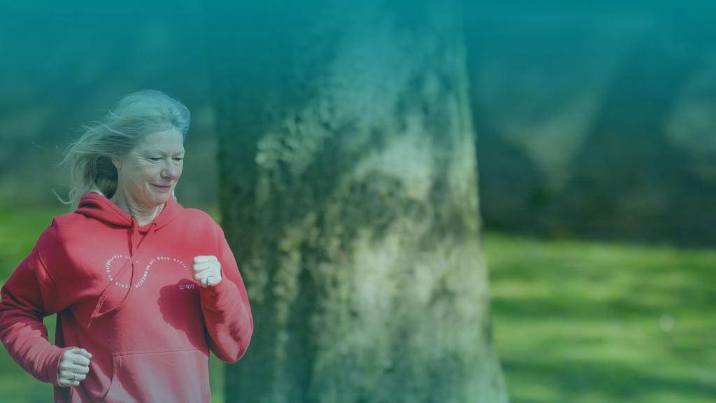 Senior woman in a red hoodie jogging in a park, with a large tree trunk and green grass in the background, under a soft blue overlay.