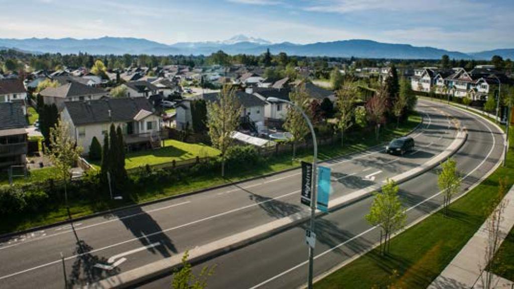 Aerial view of a suburban residential area with detached houses, a wide curved roadway with a few vehicles, sidewalks, and distant mountains under clear daylight.