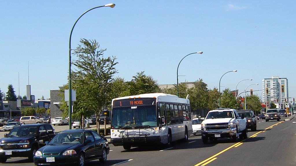 City bus travelling in traffic on a multi-lane urban street, surrounded by cars and pickup trucks on a clear day.