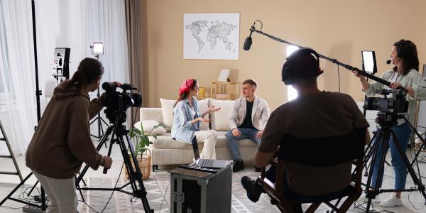 Studio video production with two people seated on a sofa in conversation, filmed by a crew using multiple cameras, softbox lighting, and a boom microphone.