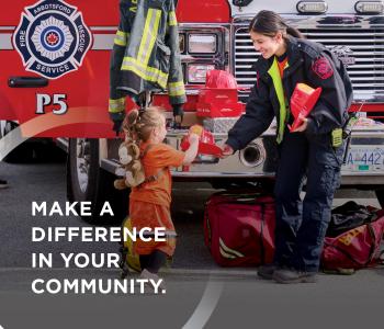 Paid On-Call firefighter at community event in front of Abbotsford Fire Truck, handing out a novelty helmet to a young child. 