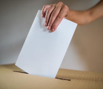 Close‑up of a hand inserting a paper ballot into a cardboard ballot box through a narrow slot.