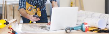 Person in tool belt using laptop and smartphone at desk with blueprints and tools, in a construction or carpentry setting.