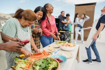 A joyful outdoor gathering with people laughing, preparing fresh vegetables, and grilling food together on a sunny day.