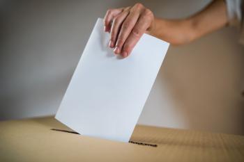 Close‑up of a hand inserting a paper ballot into a cardboard ballot box through a narrow slot.