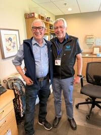 Two older adults standing together for a photo in an office, with one wearing a black Abbotsford volunteer vest and identification badge. Office furniture, shelving and artwork are visible in the background.