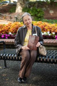 Marion Keys seated on a park bench holding a presentation folder, with colourful flower beds and trees in the background.